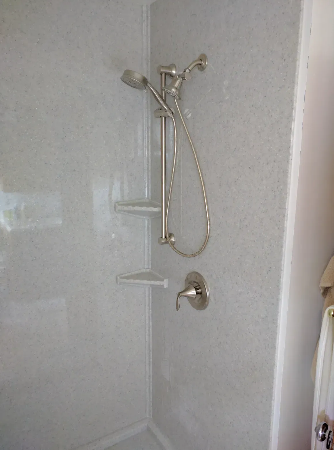 Shower stall with a light grey, speckled surround, including a handheld showerhead on a sliding bar and two corner shelves.