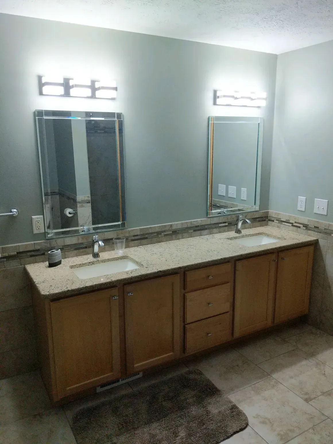 A dual-sink bathroom vanity with light wood cabinets, granite countertops, two mirrors, and two wall light fixtures.