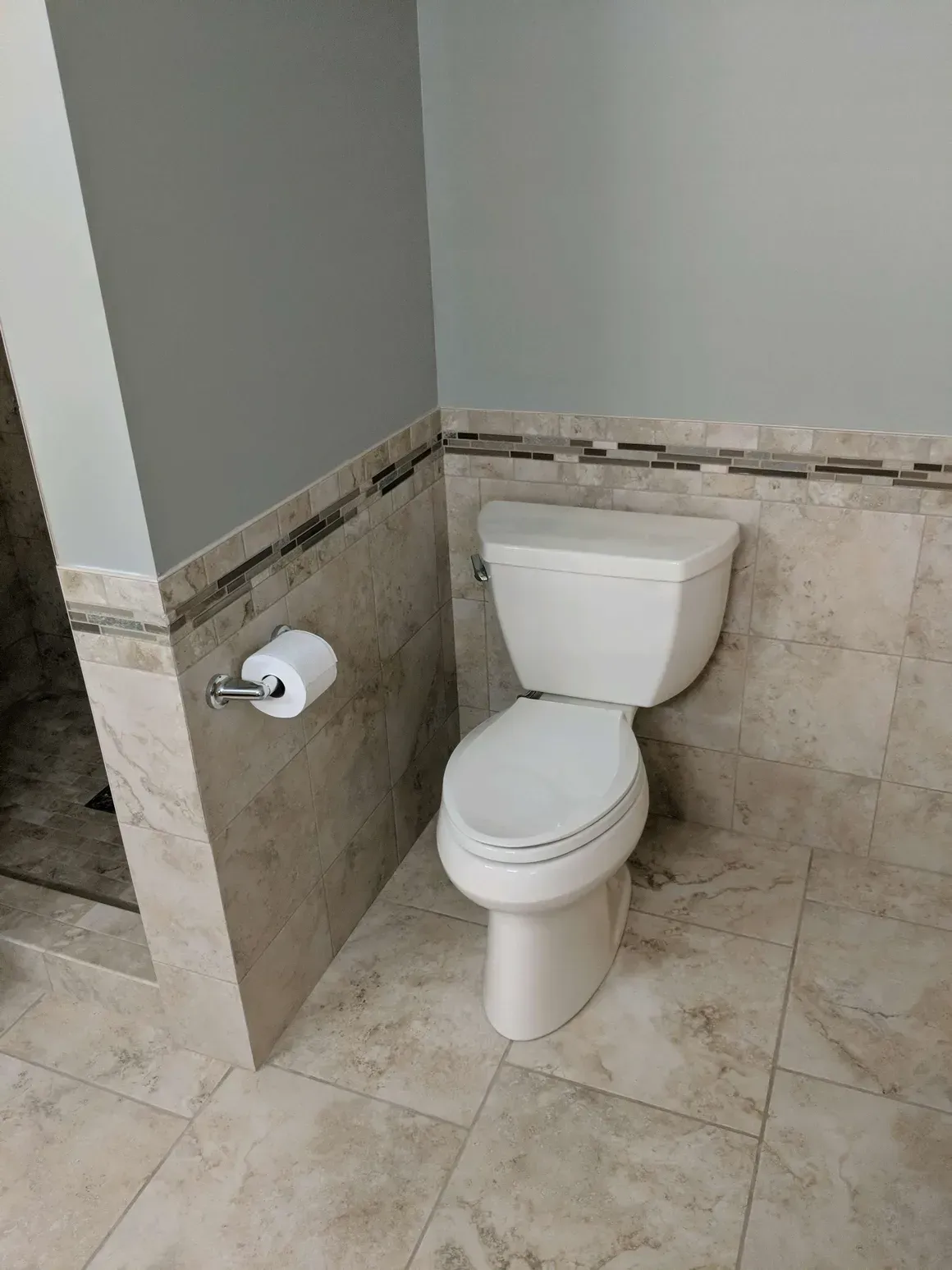 A white toilet installed in a bathroom with beige tiled walls and flooring, featuring a decorative mosaic accent tile.