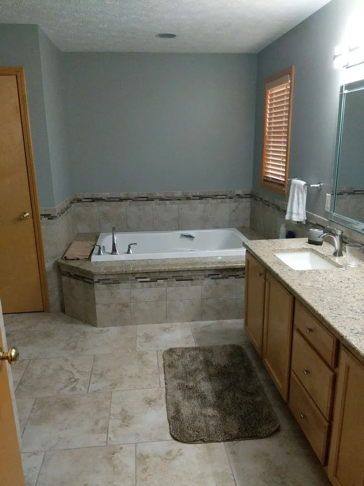A bathroom featuring a beige tiled tub surround and floor, a light wood vanity, grey walls, and a window with wood blinds.