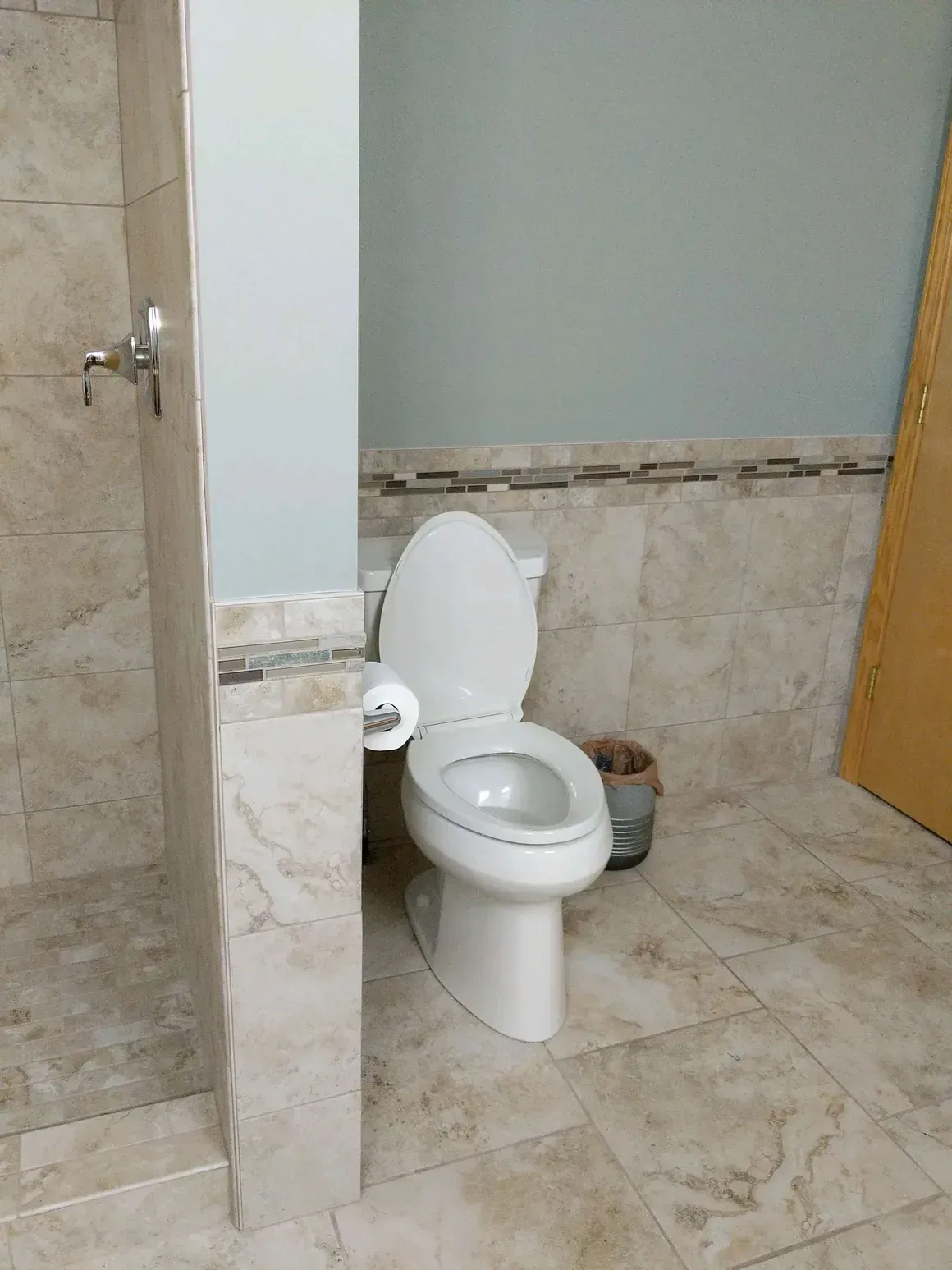 A white toilet placed in a small tiled alcove next to a shower area with a grey wall in a neutral-toned bathroom.