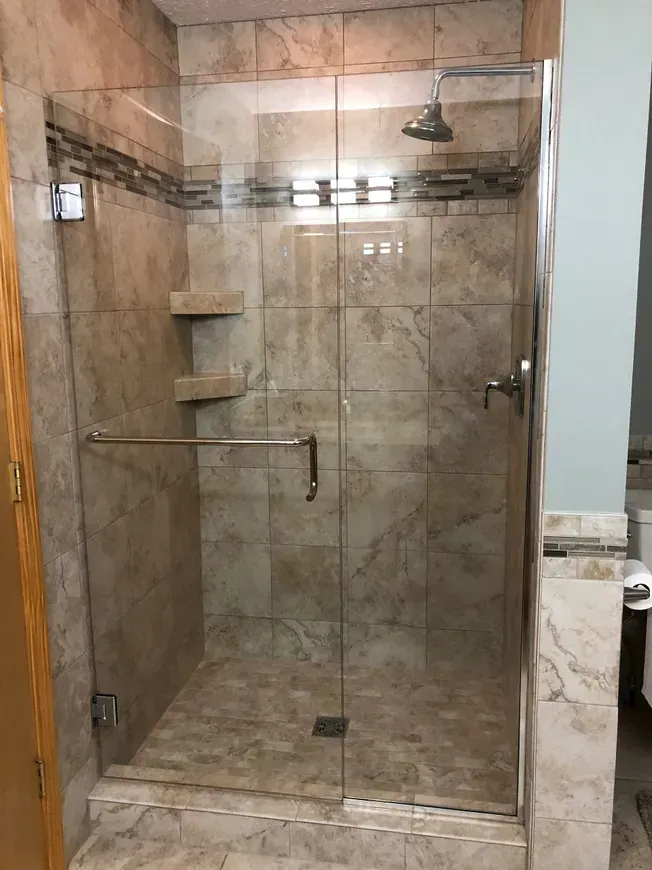 A walk-in shower with a glass door, tan stone-patterned tile walls, built-in corner shelves, and a chrome shower head.