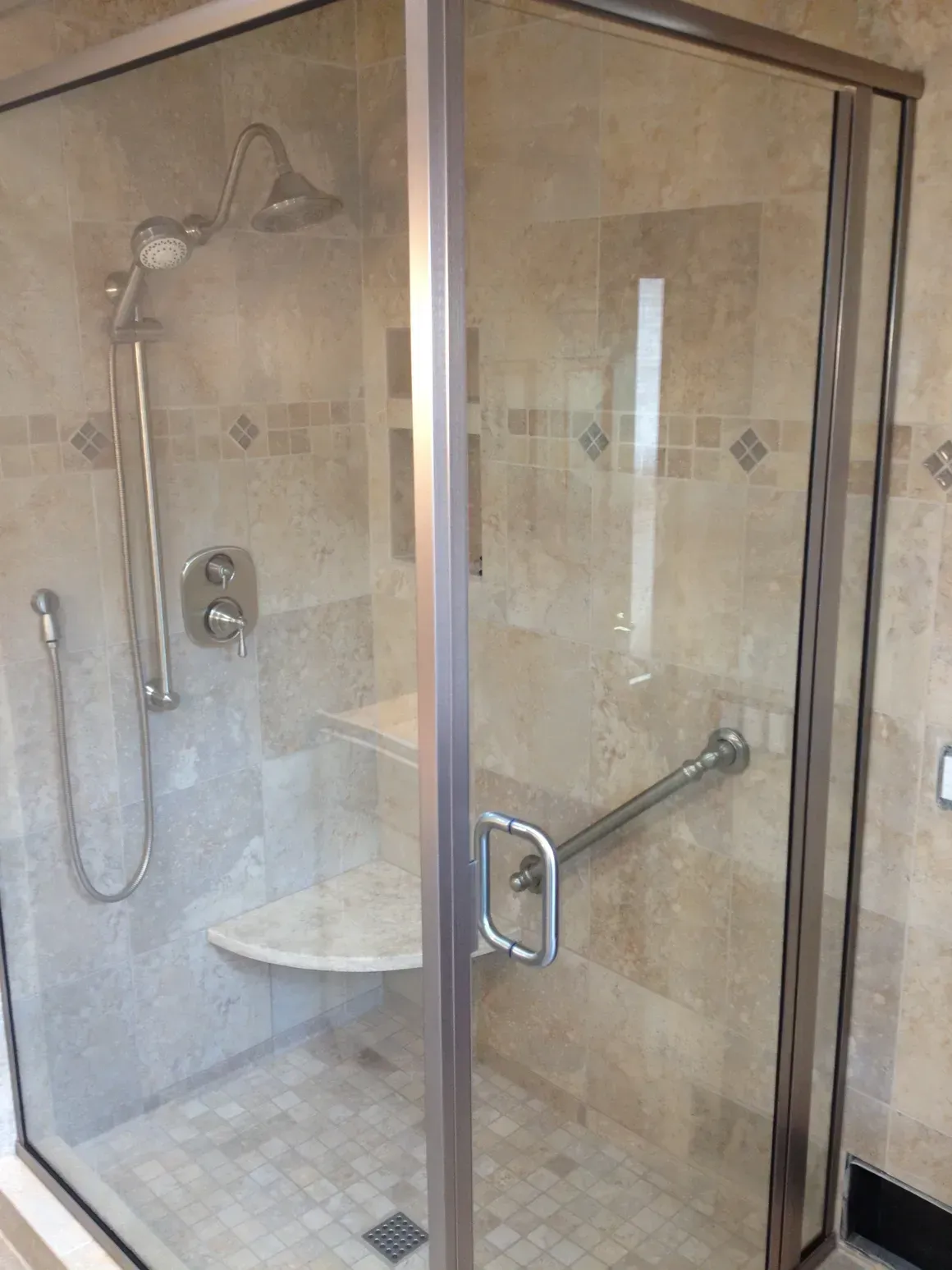 A glass-enclosed shower with beige tile walls, a built-in stone bench, a handheld showerhead, and a grab bar on the door.