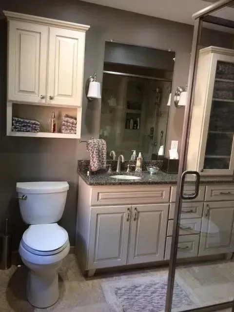 A bathroom with cream-colored vanity and wall cabinets, a granite countertop, a mirror, and a white toilet.