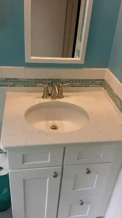 A white bathroom vanity with a cream countertop, oval sink, brass faucet, and a mirror against light blue walls.