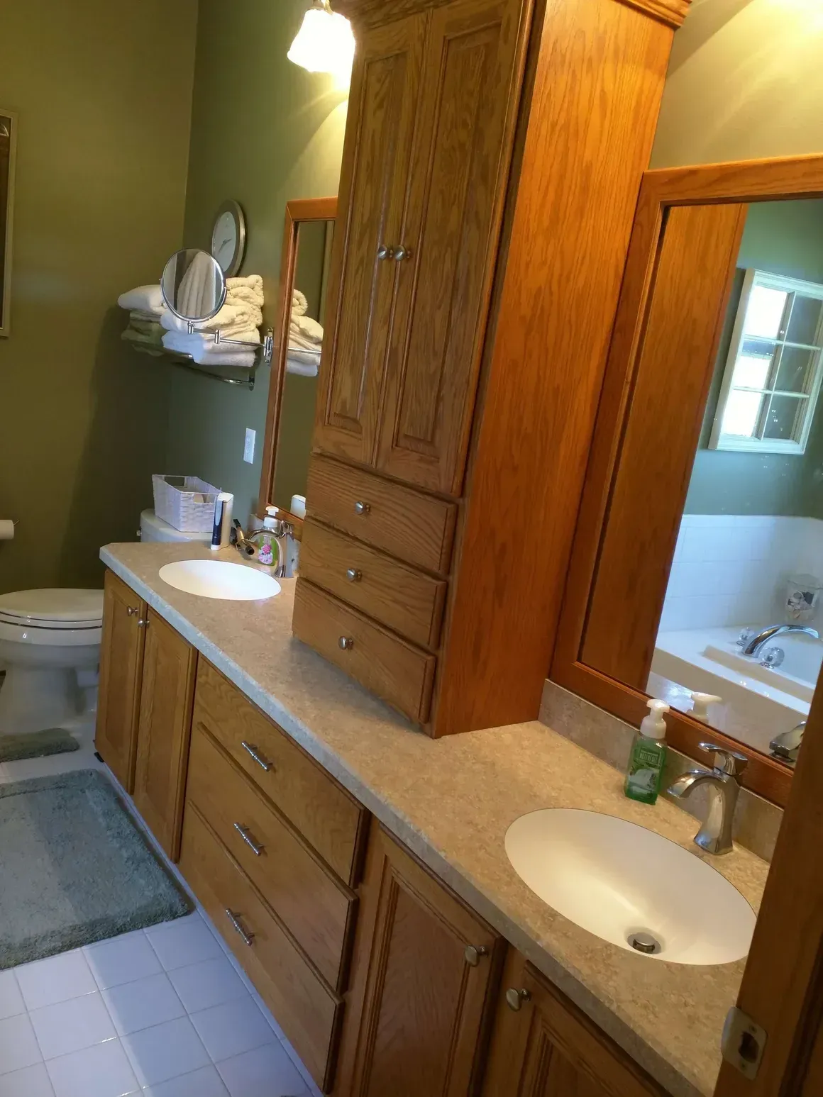 A bathroom vanity with oak cabinets, two white sinks, and a central tower, set against olive green walls.
