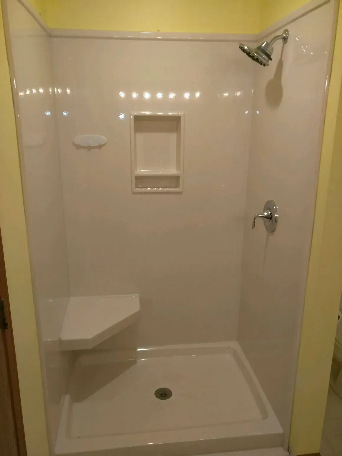 A white shower stall with a built-in corner seat, a recessed soap niche, and a chrome shower head and handle.