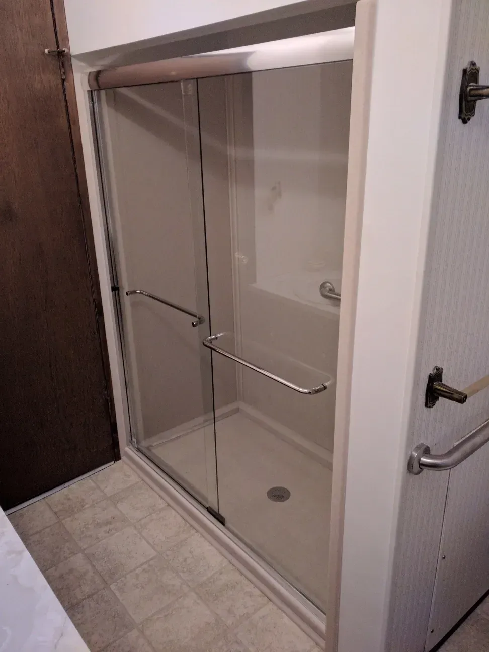 A modern glass shower enclosure with chrome handles installed in a beige-tiled bathroom with grab bars nearby.