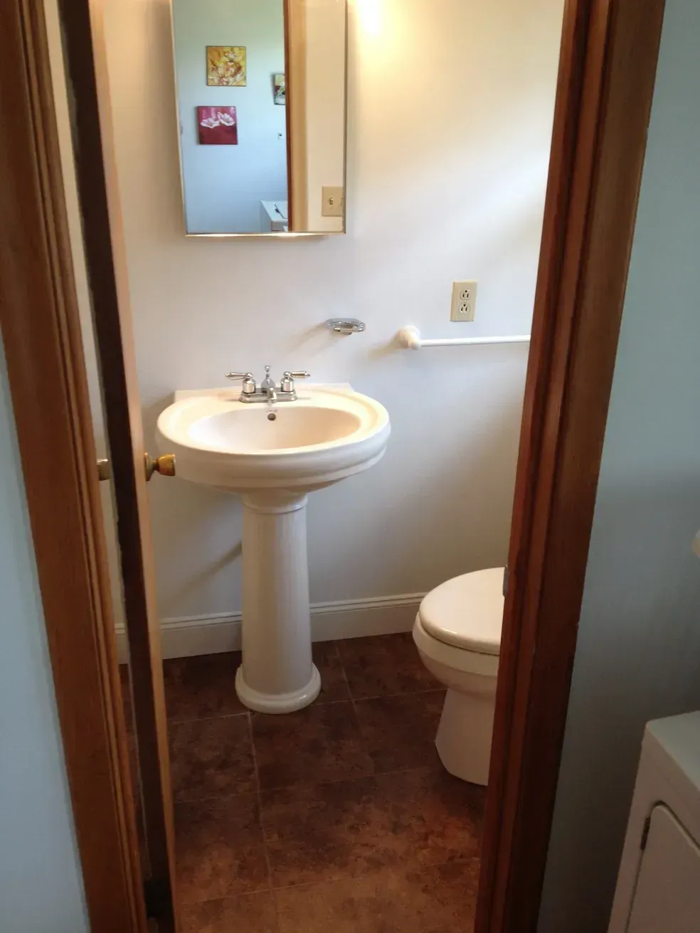 A small, neutral-toned bathroom featuring a white pedestal sink, a toilet, and a wall-mounted mirror.