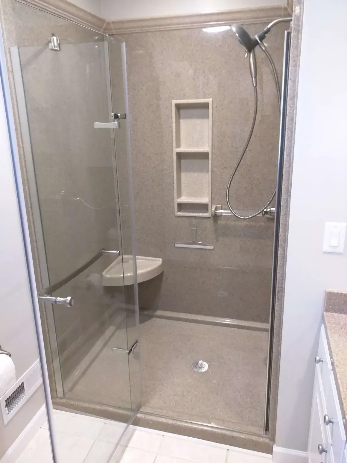A beige walk-in shower with a glass door, built-in shelving, corner seat, and adjustable handheld shower head.