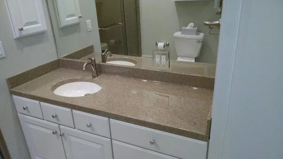 A bathroom vanity with a white cabinet, tan speckled countertop, a single sink, and a mirror reflecting the toilet.