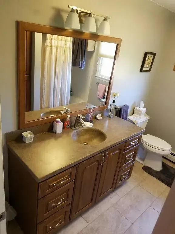 A bathroom with a wooden vanity, a brown countertop, a large framed mirror, and a toilet on tiled flooring.