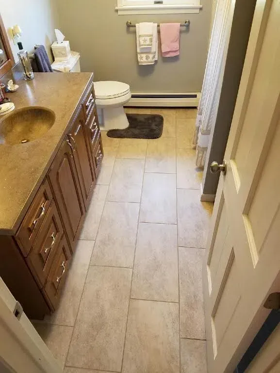 A bathroom with a wooden vanity, a beige sink, light tiled flooring, a white toilet, and towels hanging on a wall rack.