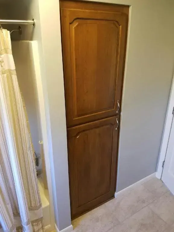 A narrow, two-door wooden cabinet set into a bathroom wall, positioned between a shower curtain and a white door.