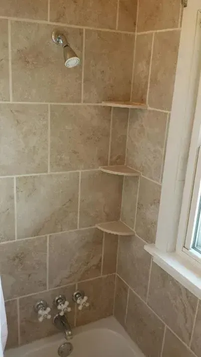A shower with tan stone-look tiles, a chrome shower head and faucet, and three built-in triangular corner shelves.