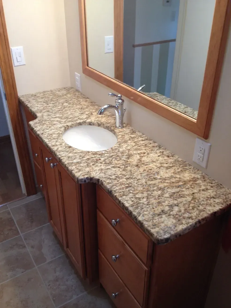 A bathroom vanity with a beige granite countertop, wood cabinets, a silver faucet, and a wall-mounted wood-framed mirror.