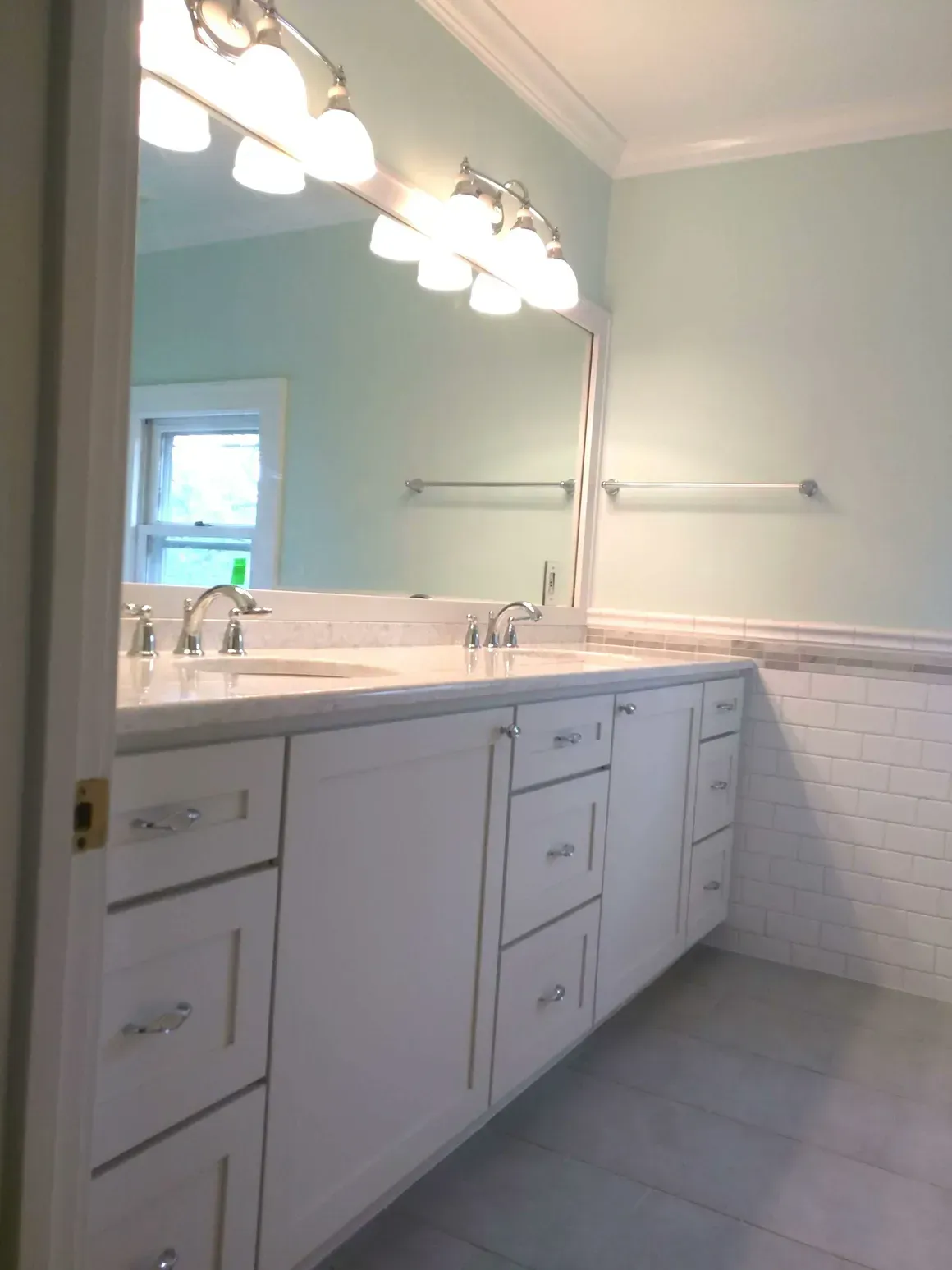 A white bathroom vanity with a large mirror and bright lighting against light green walls and white subway tile wainscoting.