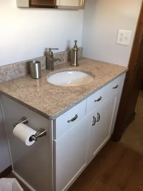 A white bathroom vanity with a beige speckled countertop, a single-handle faucet, a white sink, and a toilet paper holder.