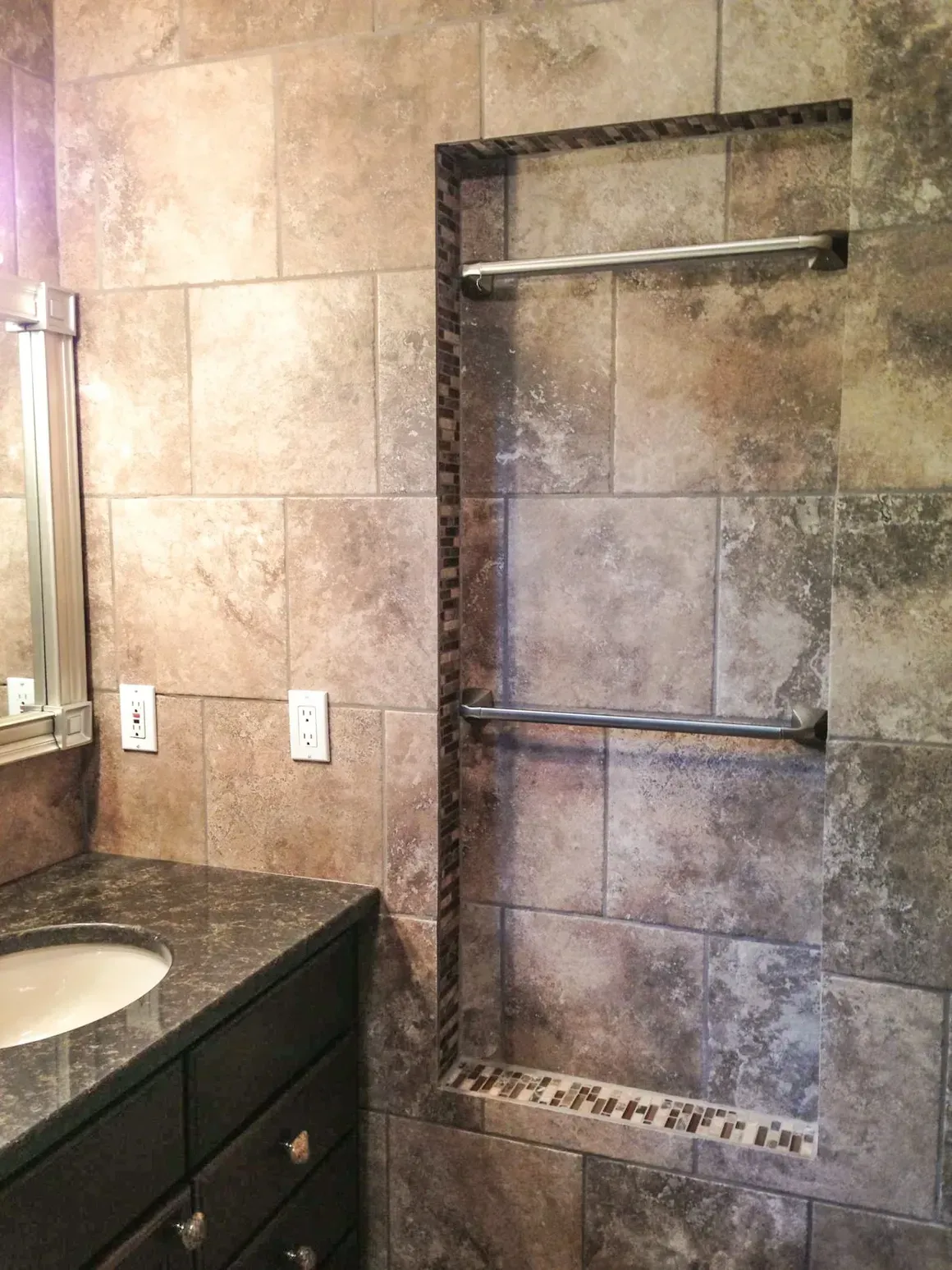 A stone-tiled bathroom wall with a vanity sink on the left and a recessed wall niche featuring two towel bars.