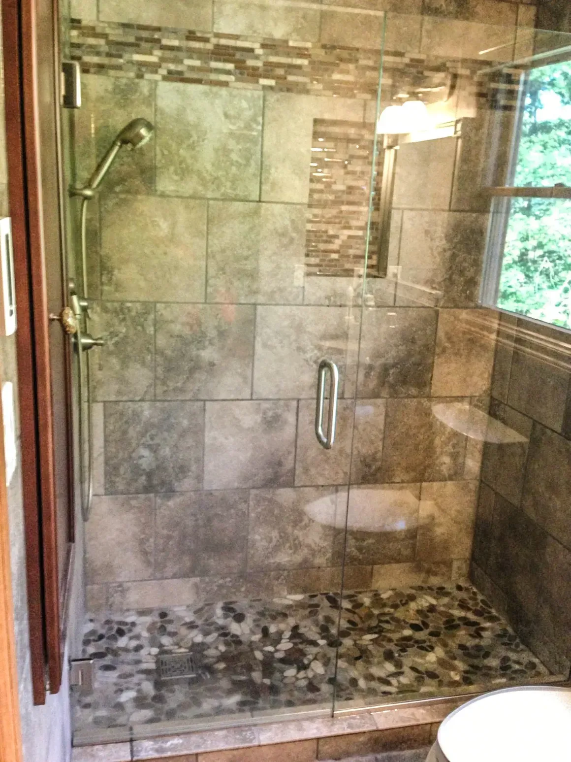 A walk-in shower with stone tile walls, a pebble floor, and a frameless glass door with a metal handle.