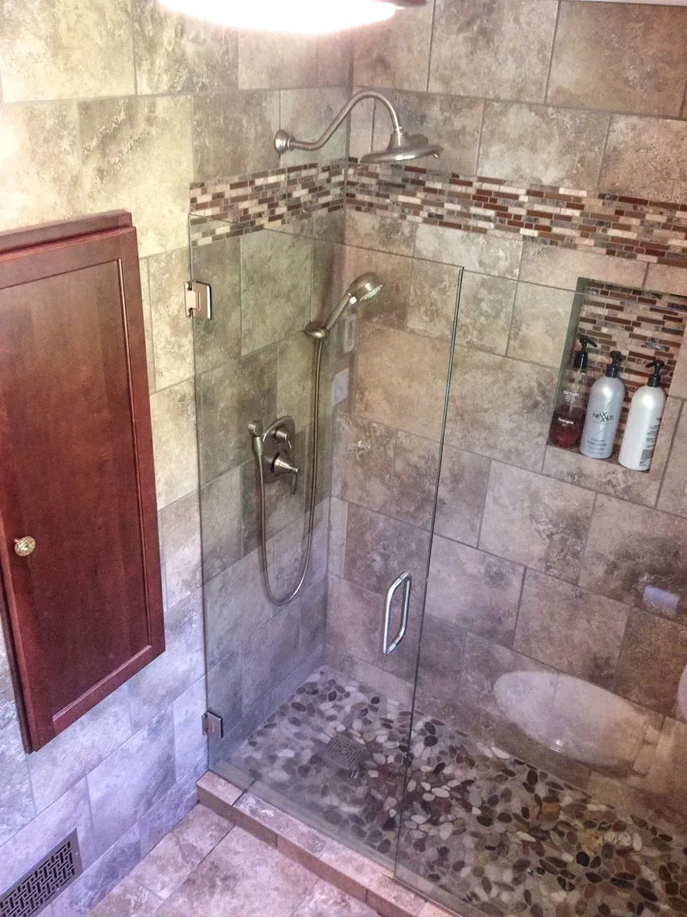 A modern shower with tan stone tile, a glass door, a pebble floor, a built-in shelf with bottles, and silver fixtures.