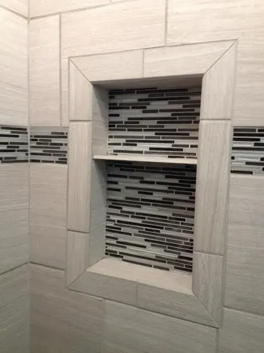 A recessed shower wall niche framed with beige tile, featuring a horizontal shelf and a decorative dark striped inlay.