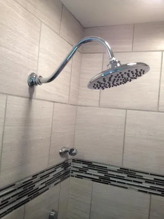 A chrome showerhead extends from a light-tiled wall with a decorative mosaic border and a small metal control knob below.