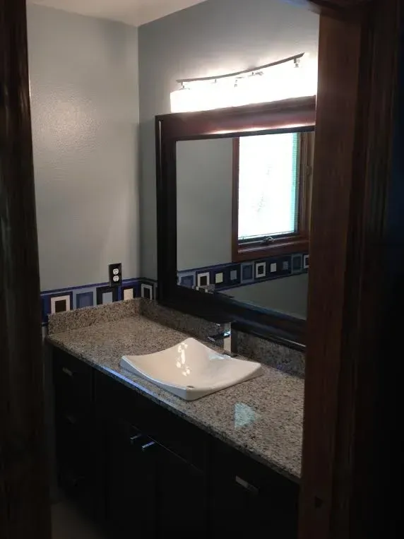 Bathroom vanity with dark cabinets, granite countertop, square vessel sink, and a framed mirror under a light fixture.