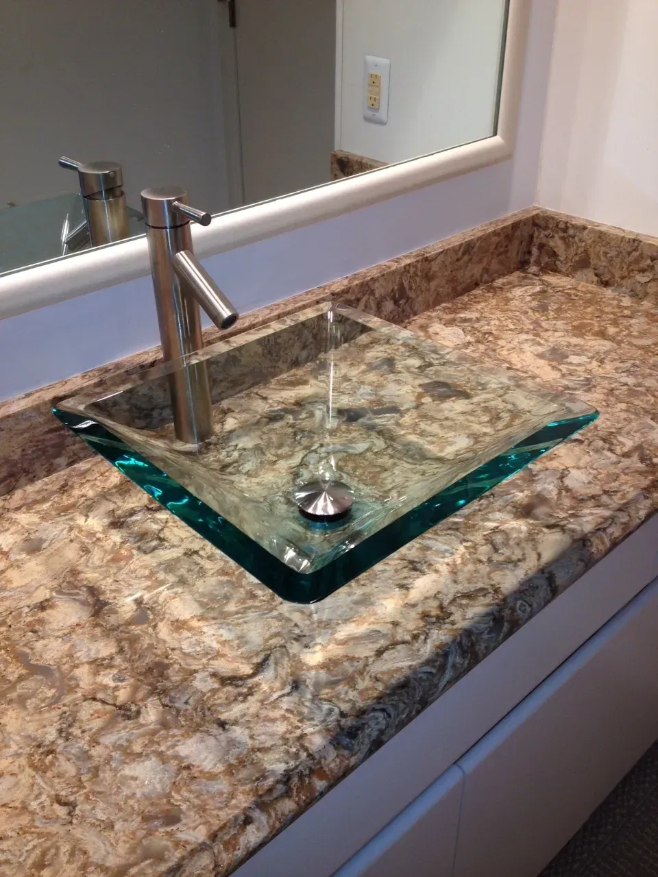 A square, clear glass vessel sink set into a speckled brown granite countertop with a tall, modern stainless steel faucet.