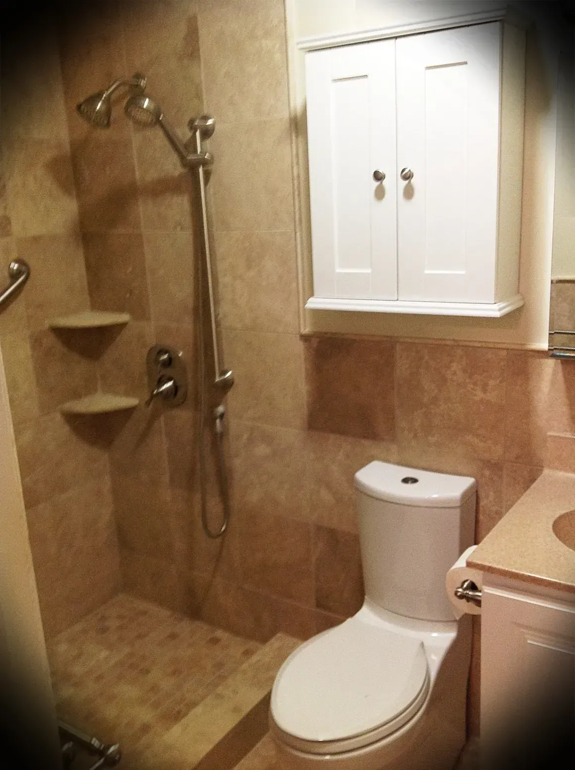 A small bathroom featuring a tan tiled walk-in shower with a dual-head shower system, a white toilet, and a wall cabinet.
