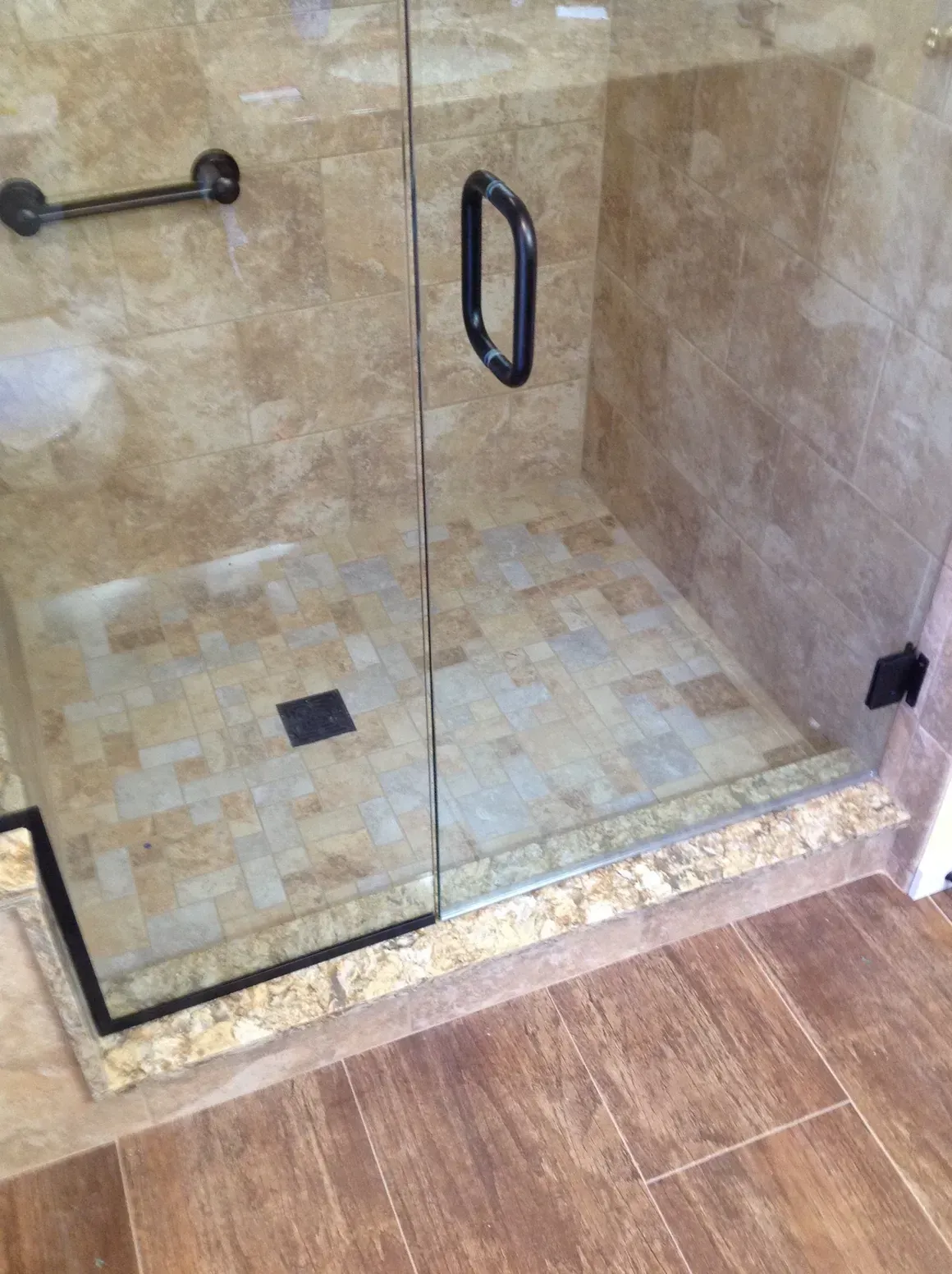 A glass shower door with a black handle, featuring beige tiled walls and a multi-toned, square-patterned tile floor.