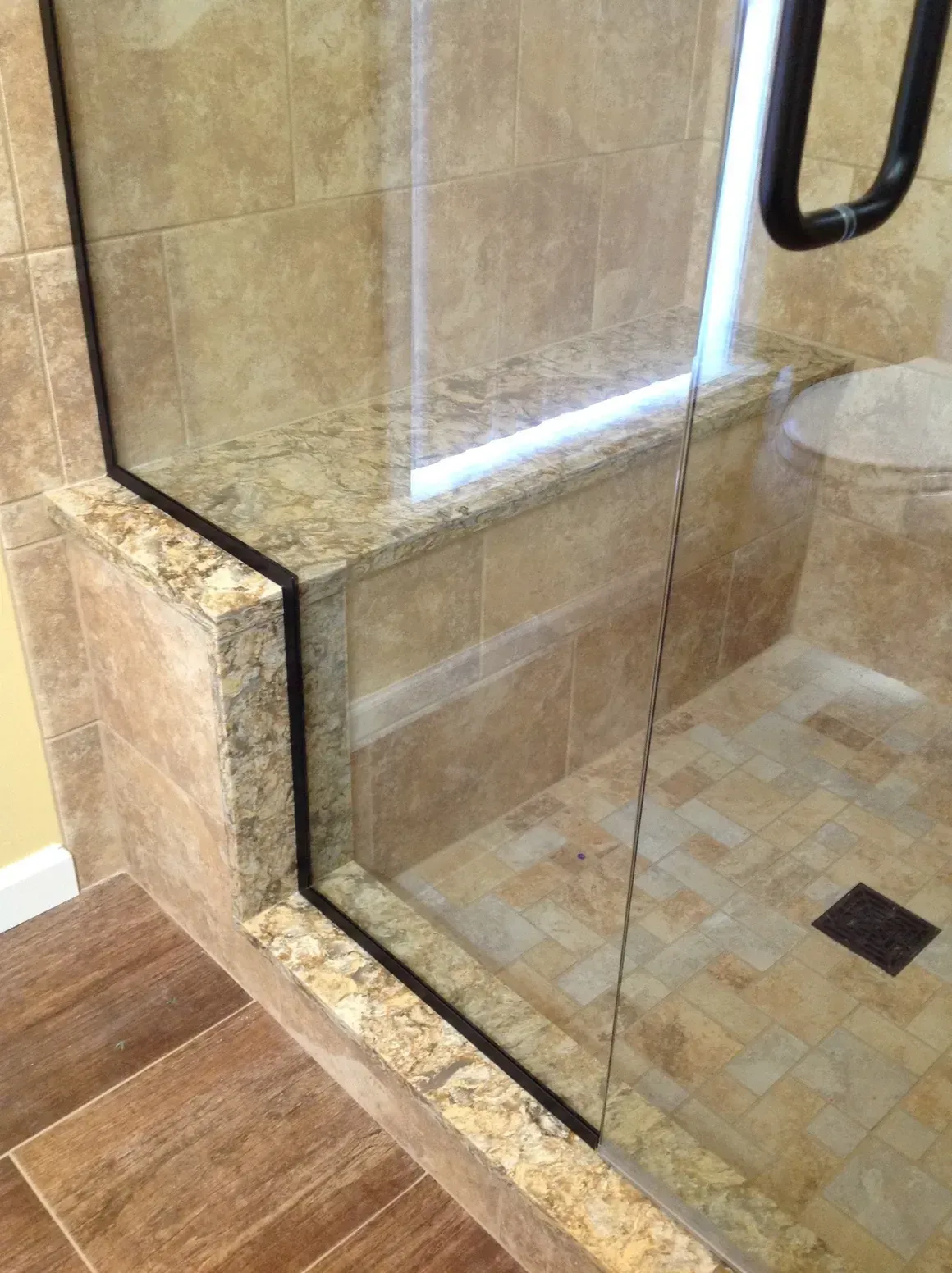 A walk-in shower with a granite bench and beige tile walls, framed by a glass door with a dark handle.