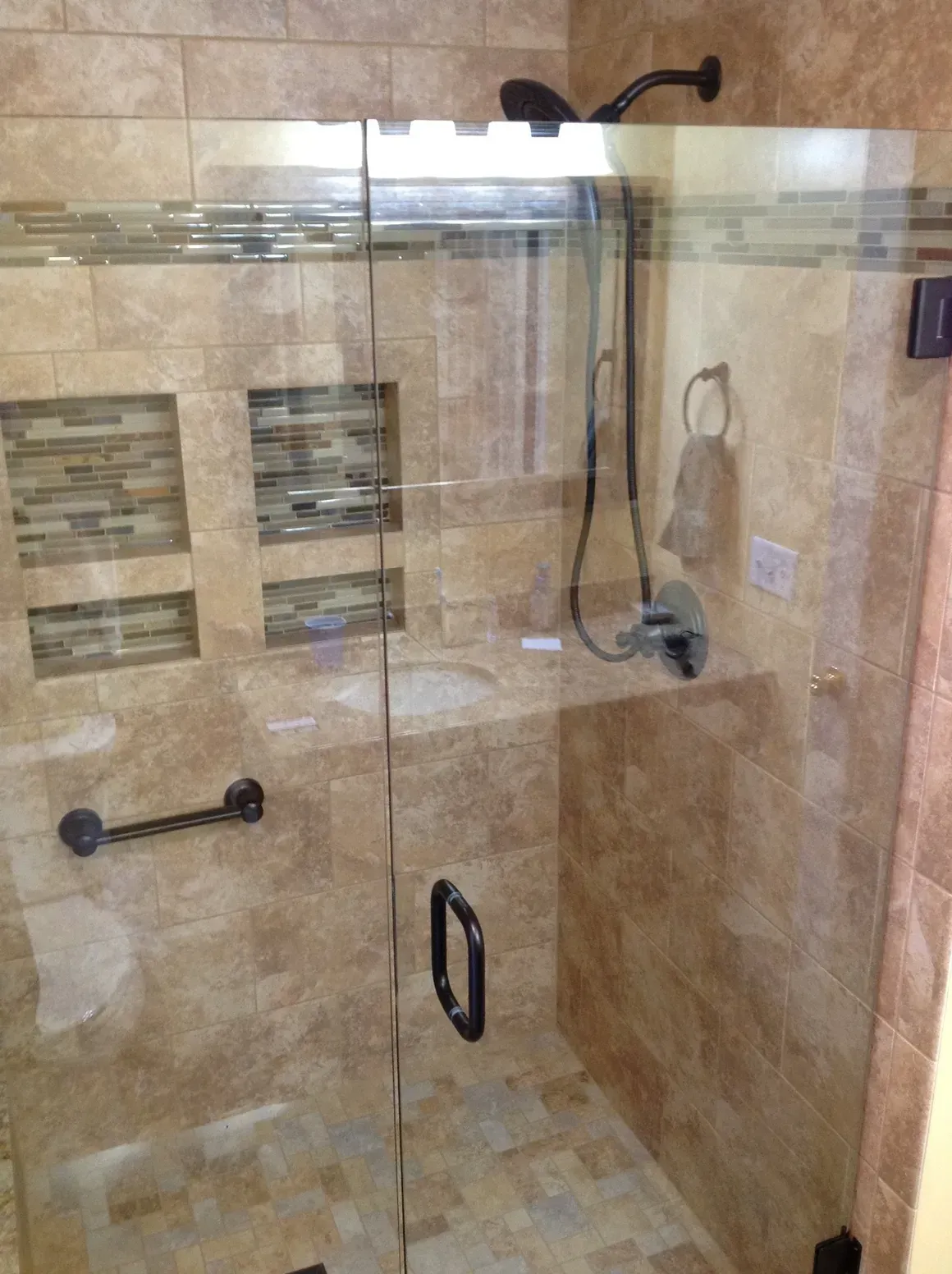 A walk-in shower with tan tiled walls, mosaic niches, a glass door, a black handle, a grab bar, and a handheld showerhead.