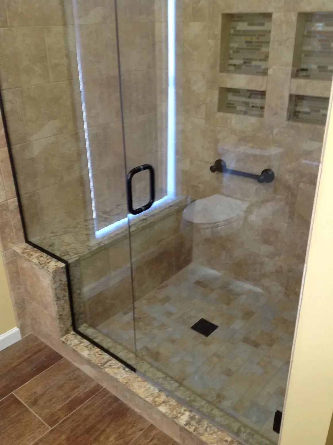 A tiled walk-in shower with a glass door, a built-in seat, a black grab bar, and two recessed shelves on the back wall.