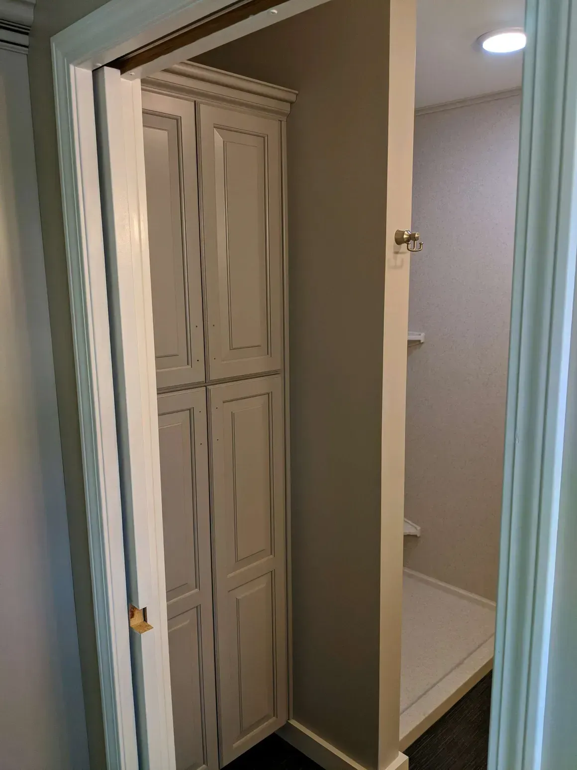 A tall, beige, four-paneled storage cabinet stands inside a room doorway next to a small, white-tiled walk-in shower.