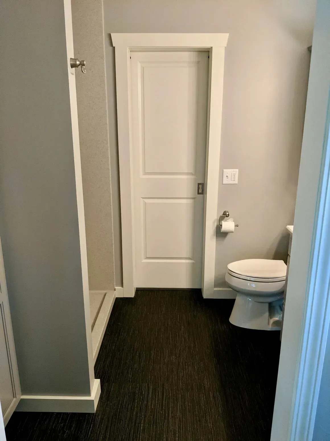 A bathroom with grey walls and dark flooring, featuring a white door, a toilet, and the edge of a shower stall.