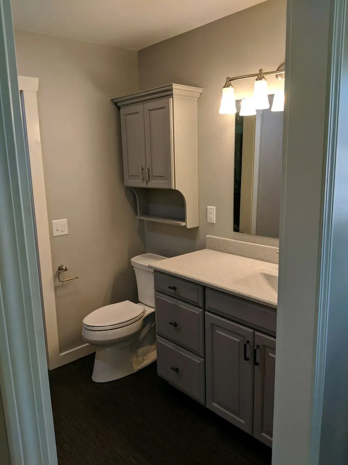 A bathroom with gray cabinetry, a white toilet, a vanity with a white countertop, and a vanity light fixture.
