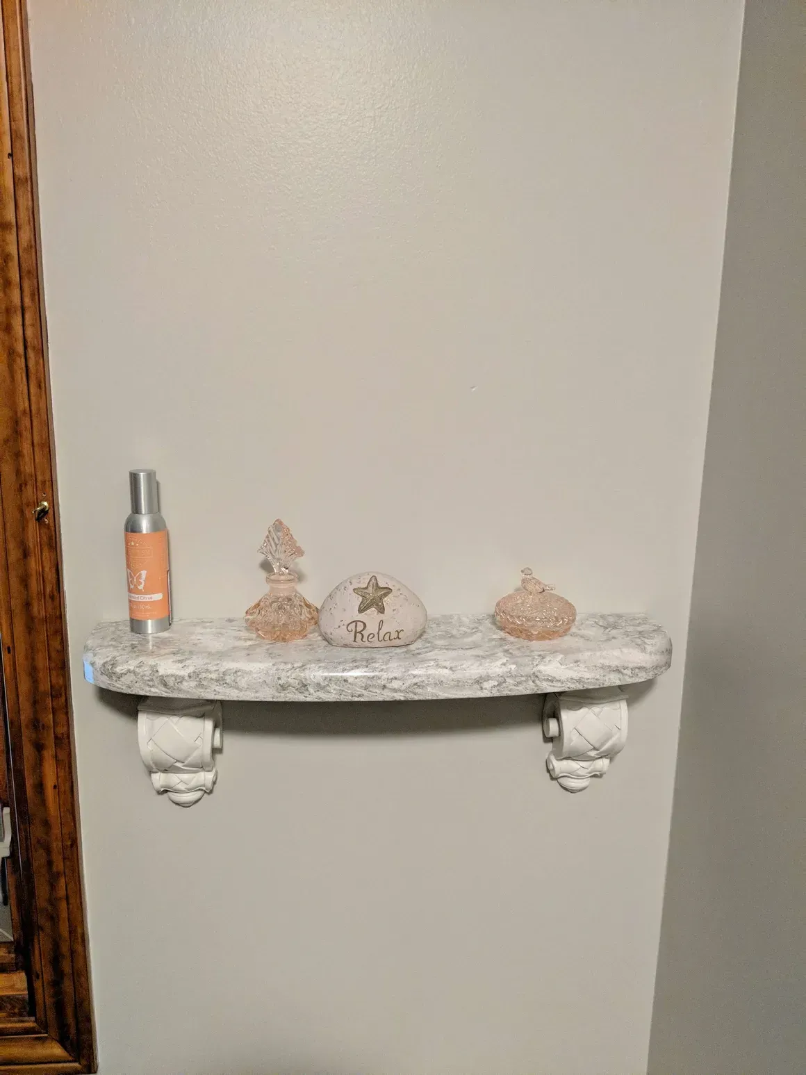 A white floating shelf with two decorative brackets, holding a spray bottle and three small decorative trinkets.
