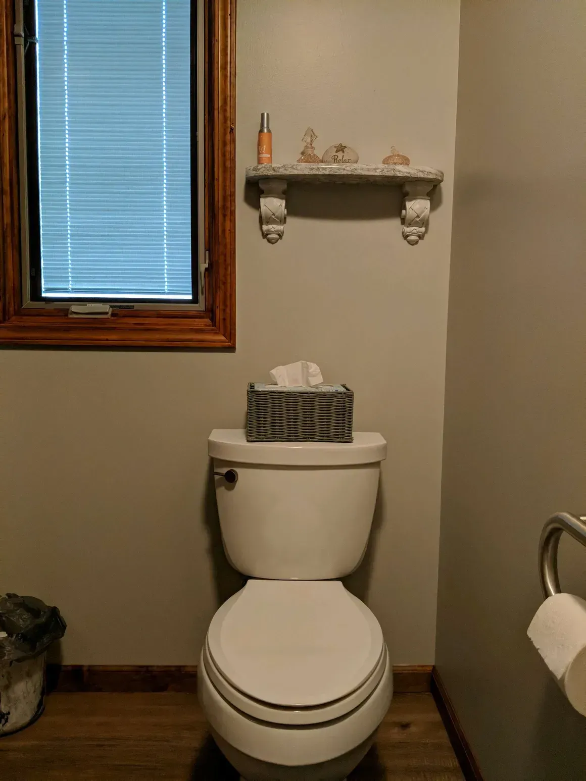 A toilet sits in a room with light grey walls, a wooden window frame, a small white shelf above, and a toilet paper holder.