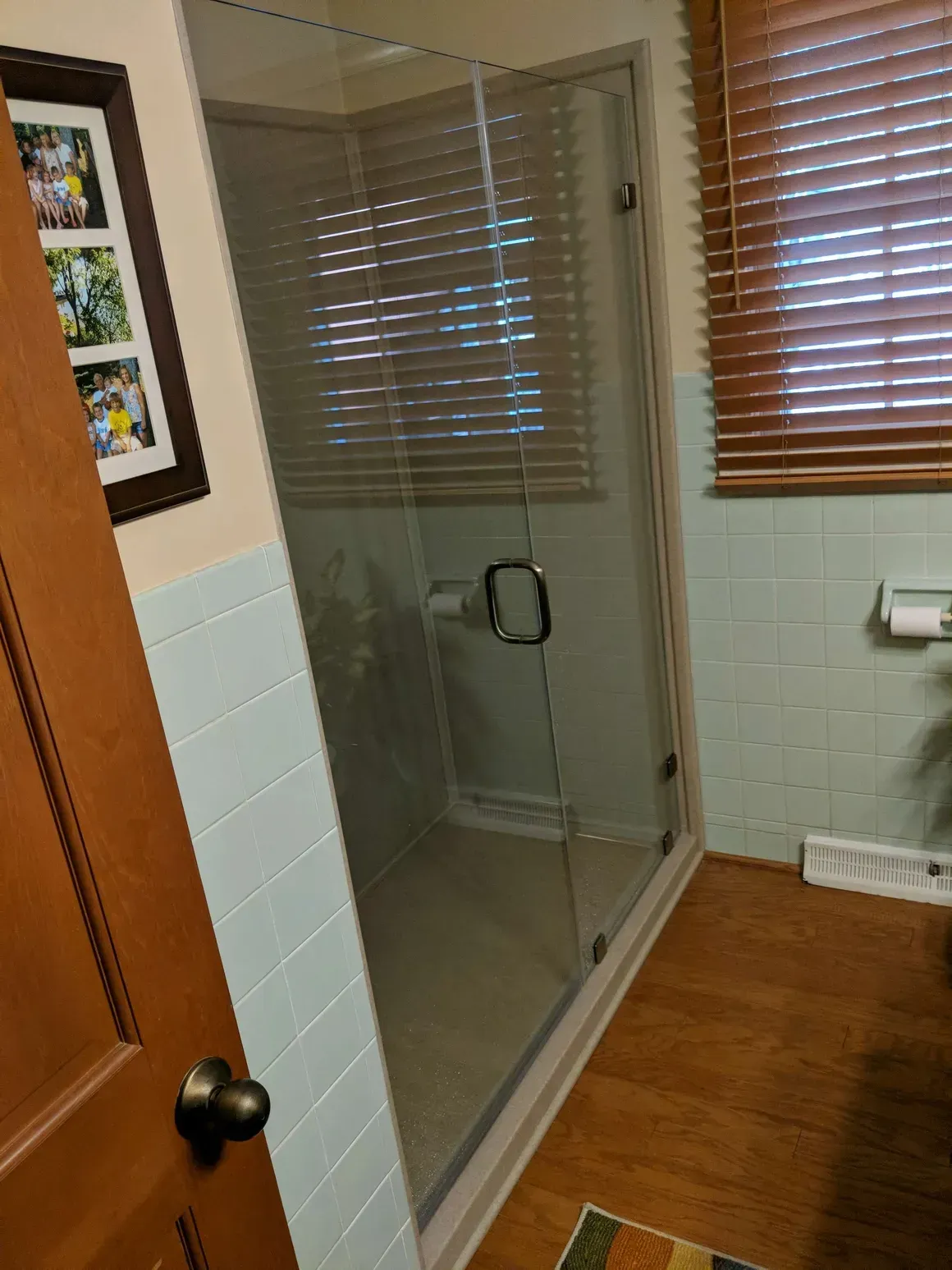 A glass-enclosed shower stall in a bathroom with light blue wall tiles, wooden blinds, and a wood-toned door.