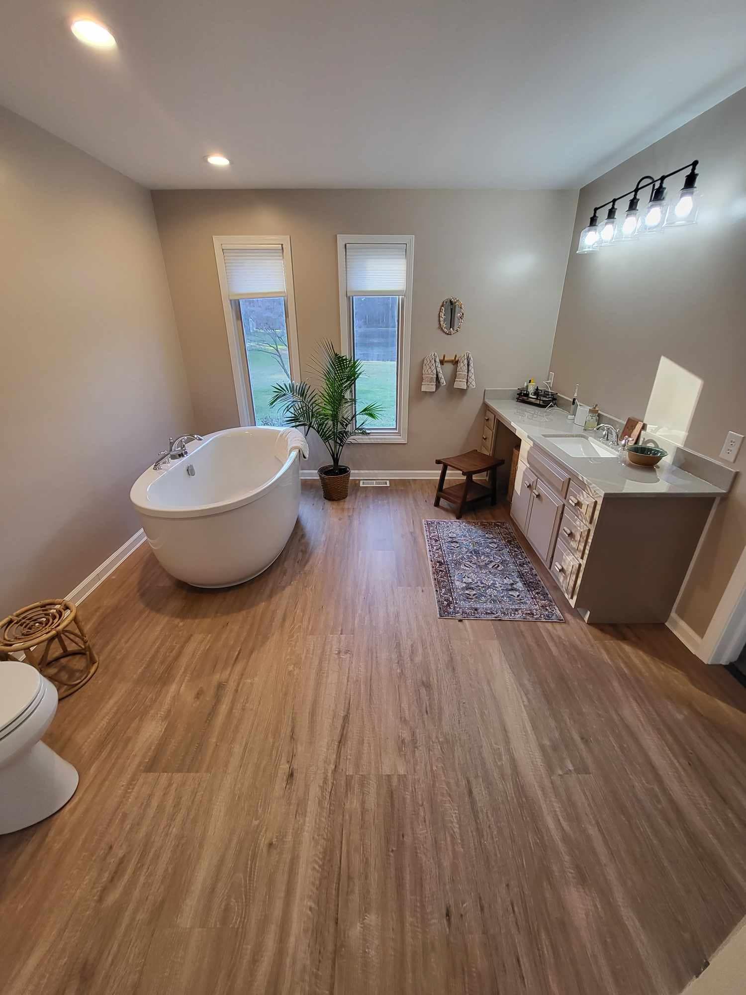 A bathroom with a freestanding white tub, two windows, a wooden stool, a patterned rug, and a gray vanity with lighting.