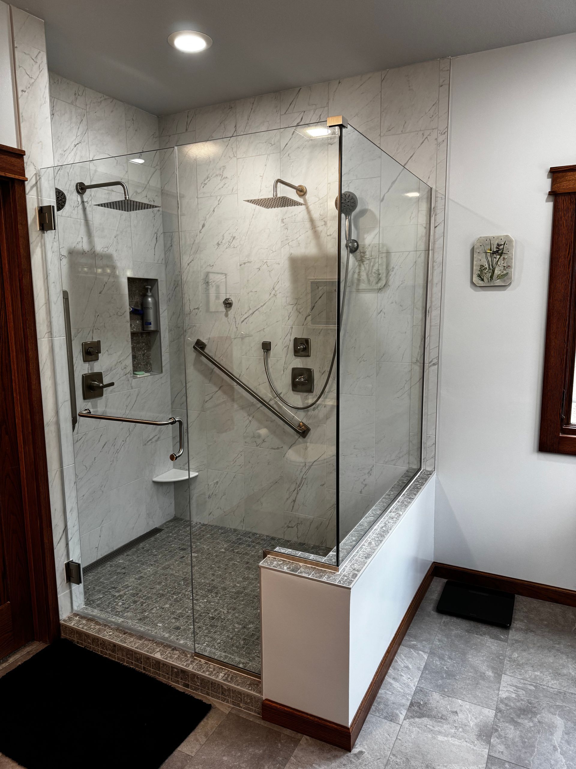 A walk-in shower with white marble walls, two showerheads, a glass enclosure, and a chrome support grab bar.