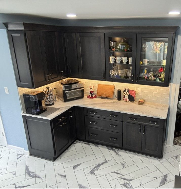 A dark kitchen corner with black cabinets, white stone countertops, herringbone floor tiles, and a light-up glass display.