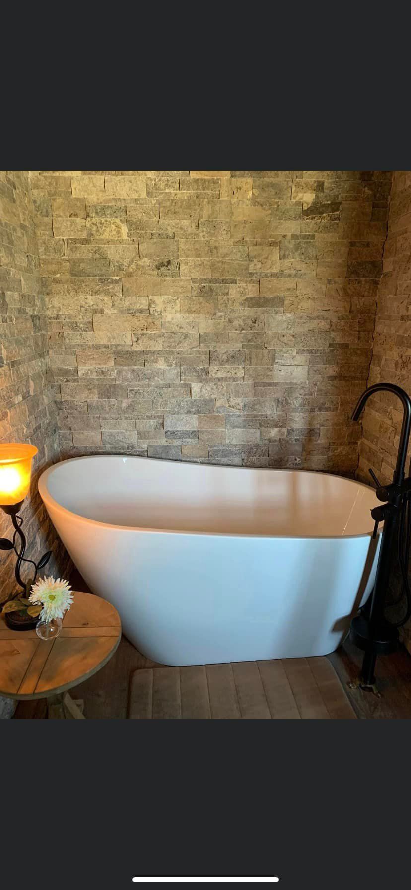 A white freestanding bathtub sits against a rustic tan stone wall next to a wooden side table with a lamp and vase.