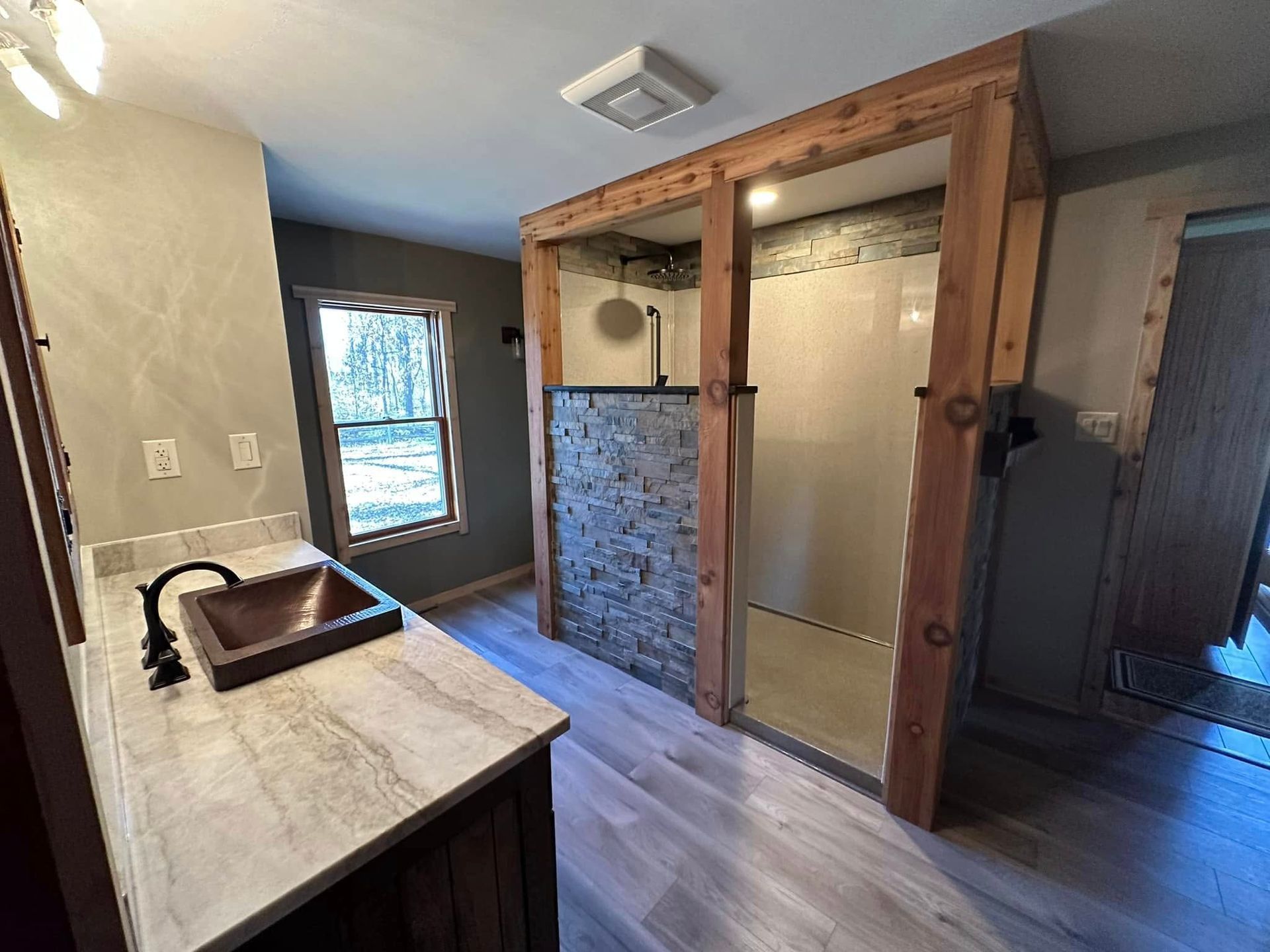 A modern bathroom featuring a stone-accented walk-in shower, a rustic vanity with a copper sink, and light wood flooring.