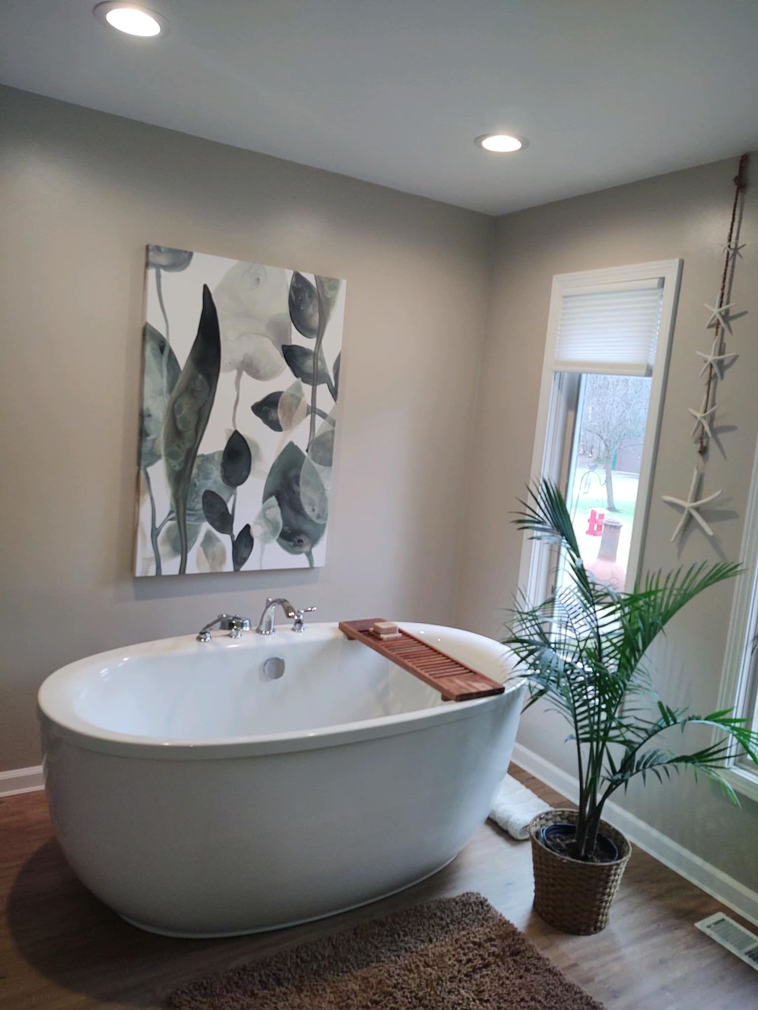 A white freestanding oval bathtub sits in a room with light walls, a large floral painting, and a potted palm tree.