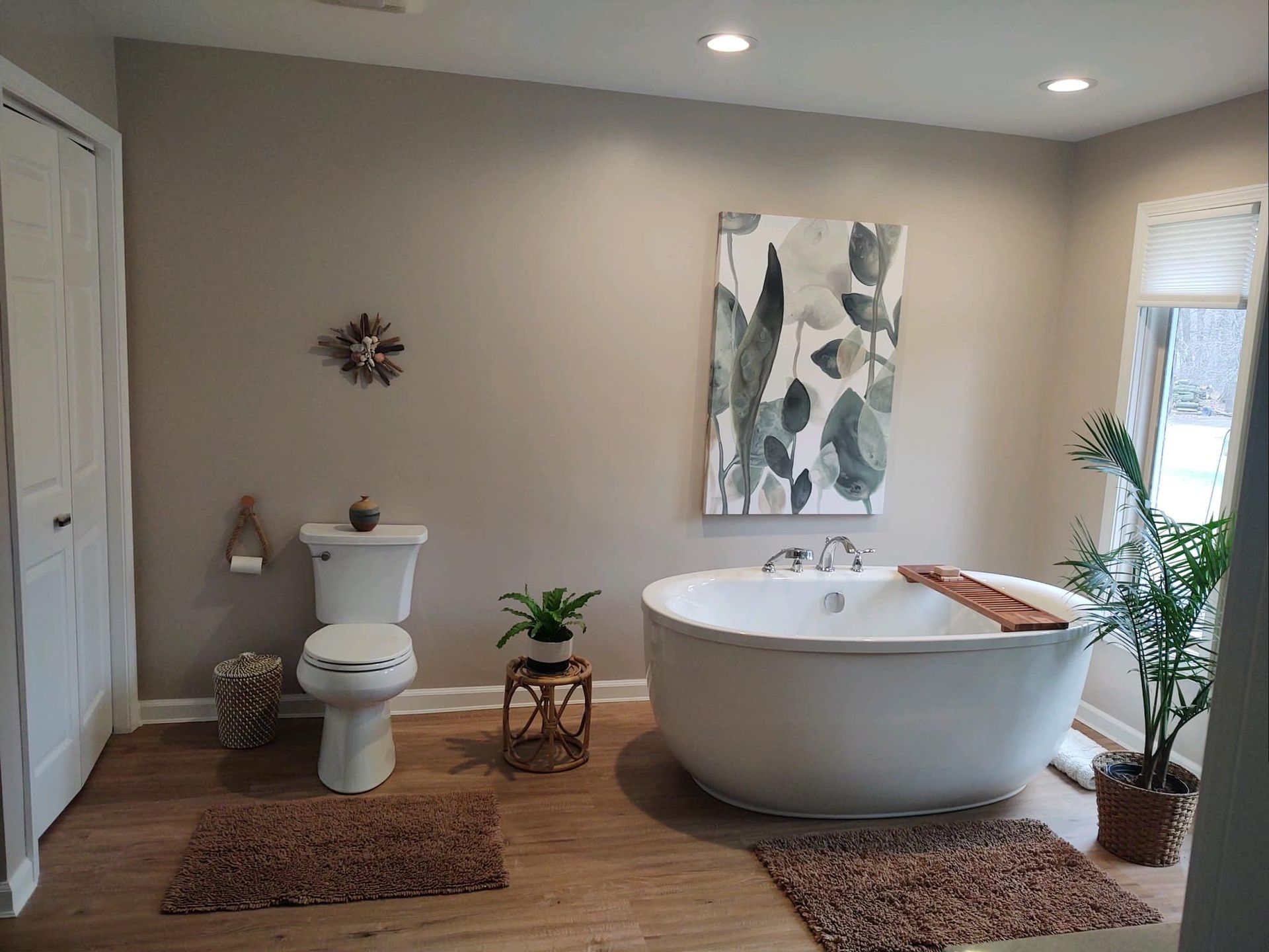 A bathroom with a white freestanding tub, a toilet, a neutral wall with floral art, a plant, and two brown rugs on floors.