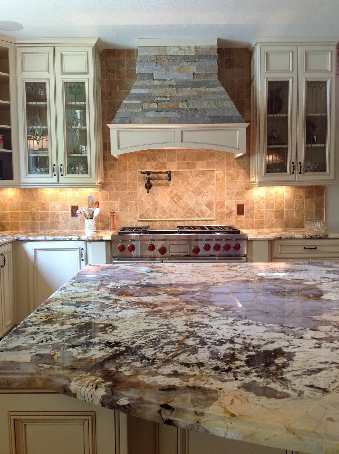 A kitchen features cream cabinets, a patterned granite island, and a stone range hood above a stainless steel stove.