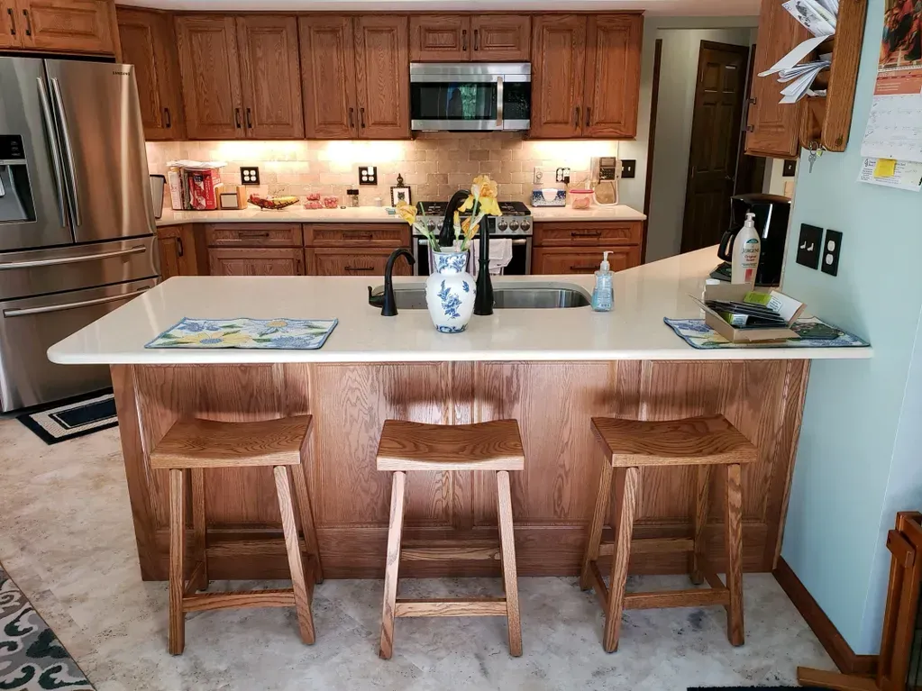 A kitchen island with three wooden bar stools in front of an L-shaped counter, oak cabinets, and stainless steel appliances.