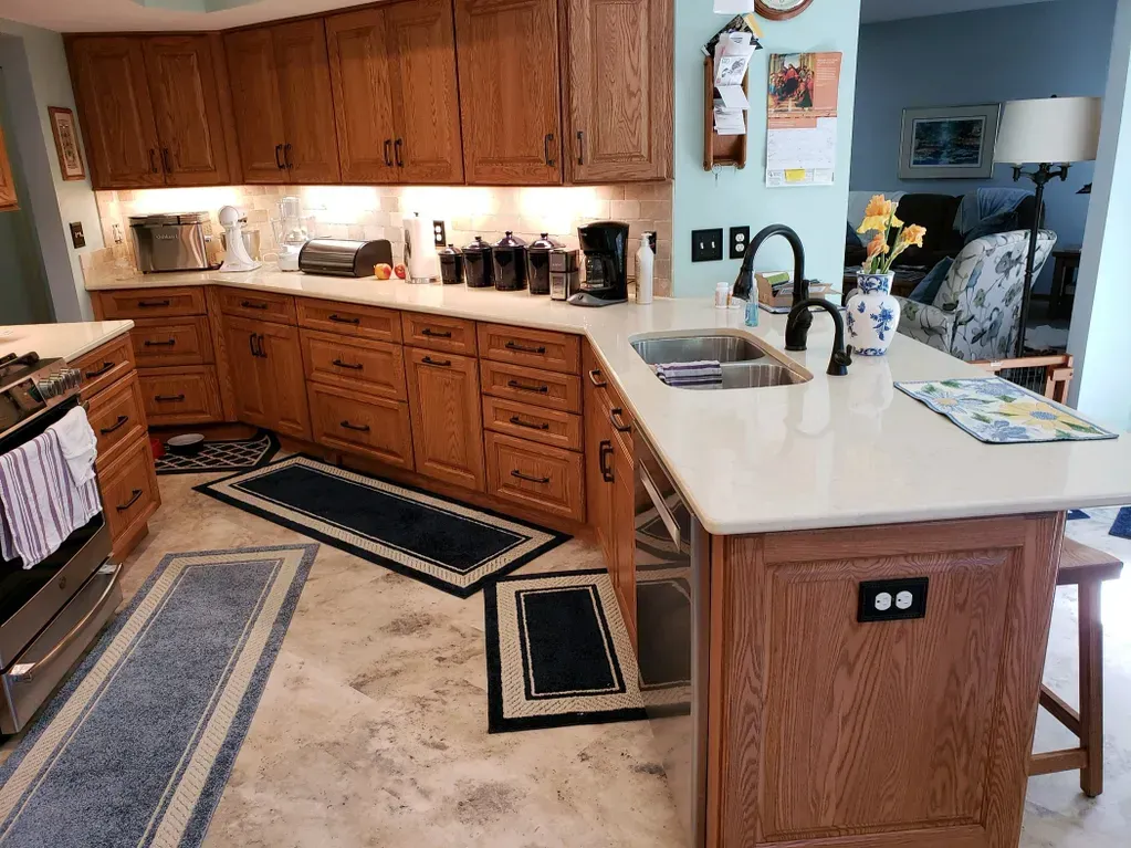 A bright kitchen with oak cabinets, white countertops, stainless steel appliances, and blue patterned floor runners.